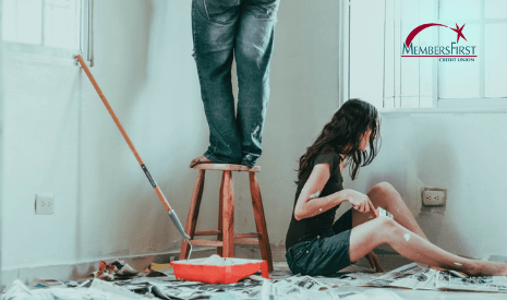 Man in baggy jeans Standing in Chair and Woman in dark shirt and jean shorts with paint on her knee Sitting in the floor in white room painting