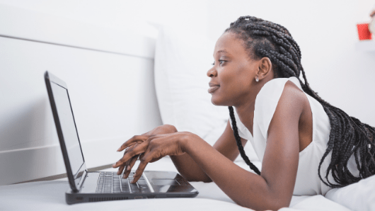 African American woman in white dress laying on bed looking at computer